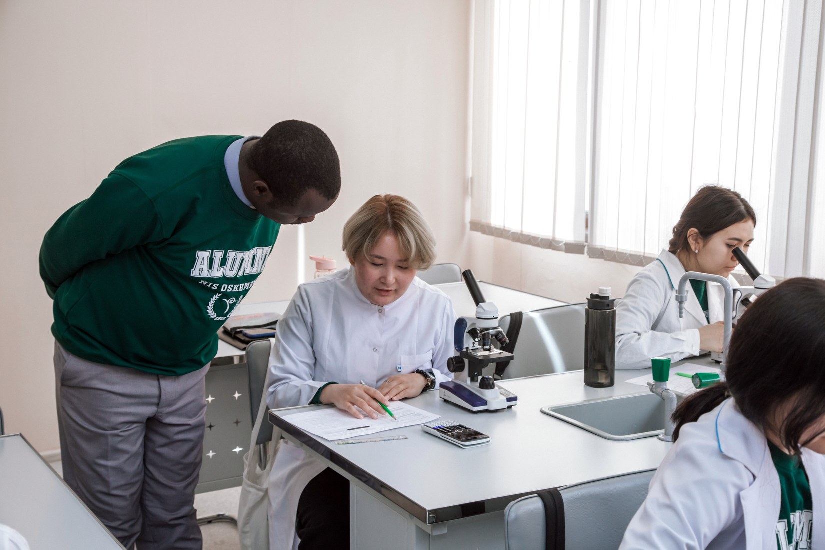 Students in white coats working with microscopes at a school laboratory bench, one of them in an NIS Oskemen alumni sweatshirt.