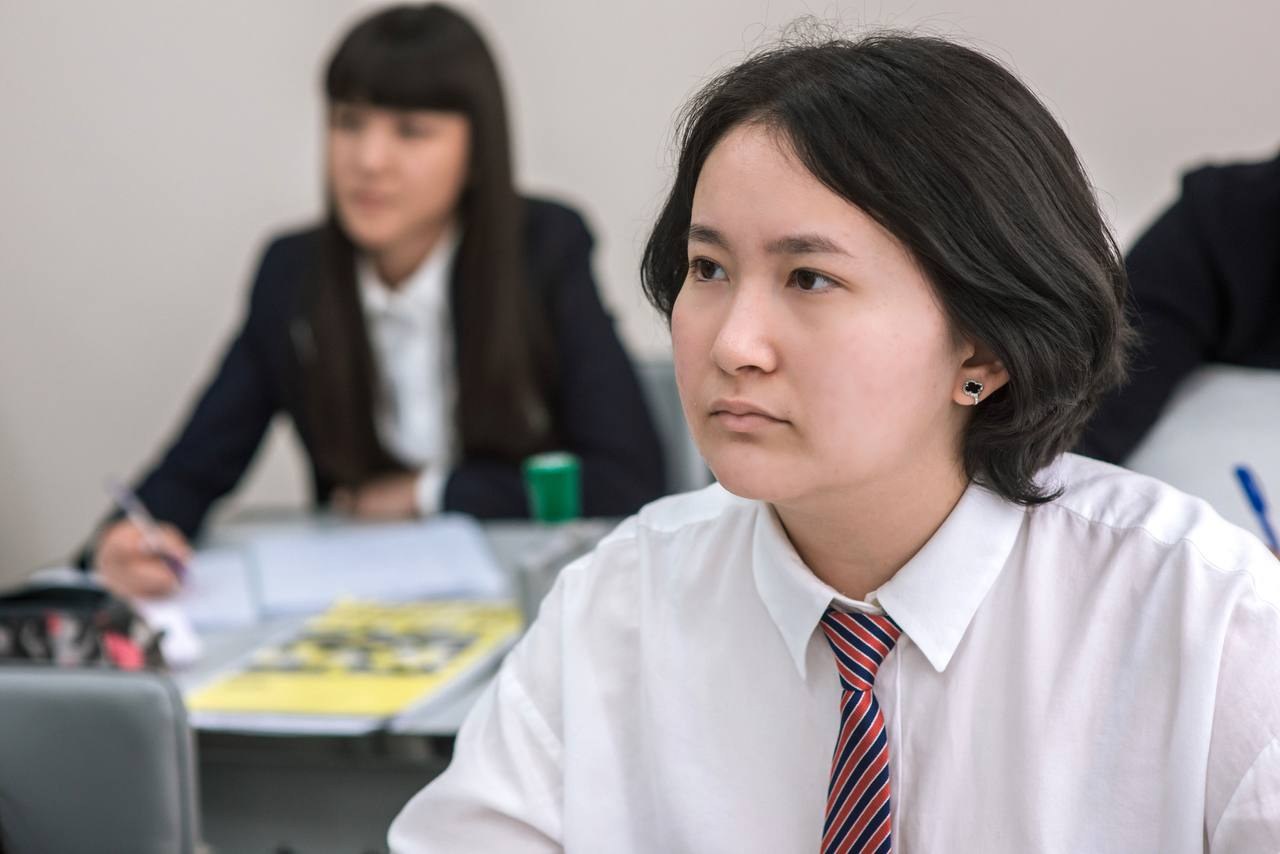 A student in school uniform at a classroom desk, looking towards the front of the room.