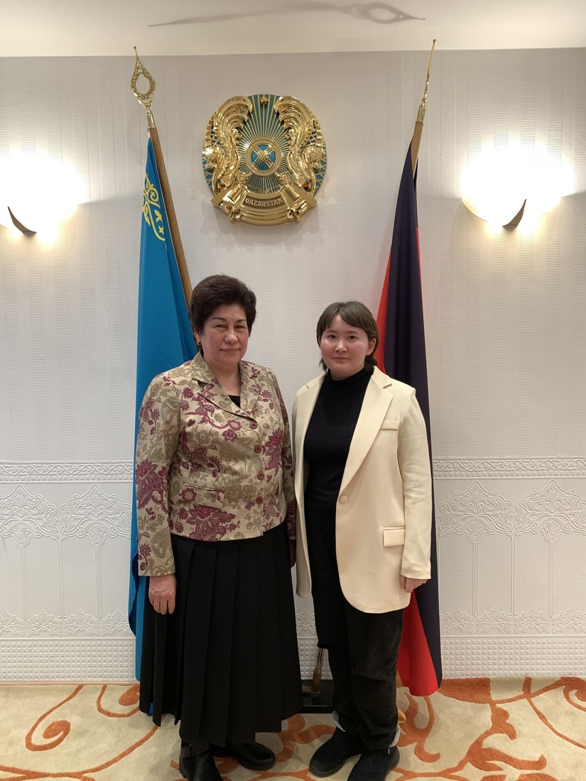 Two women standing in front of the Kazakhstani state emblem and two flags in an embassy reception room.